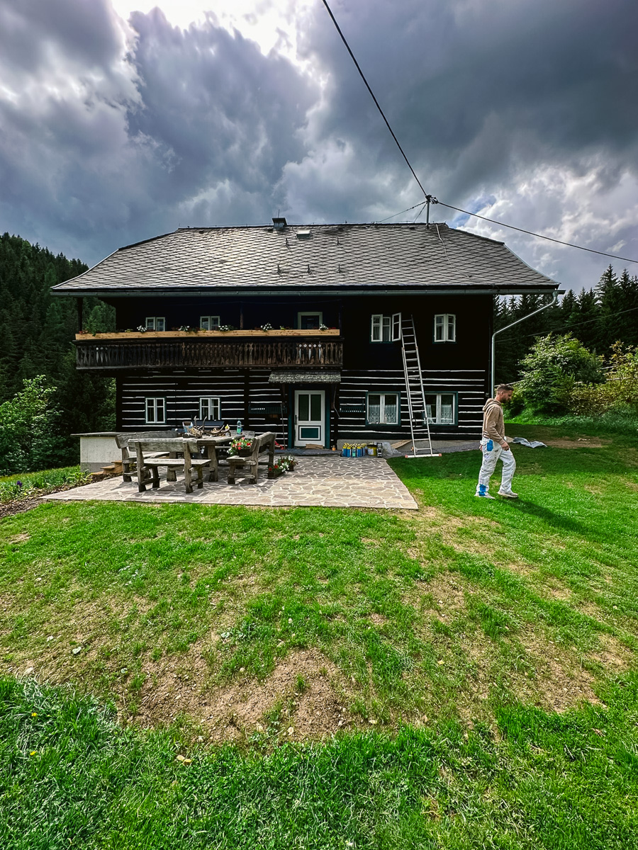 Traditionelles Holzhaus in den Bergen; Maler saniert dunkle Holzfassade mit Leiter und Balkon – Außenanstrich/Lasur vor Gewitterhimmel