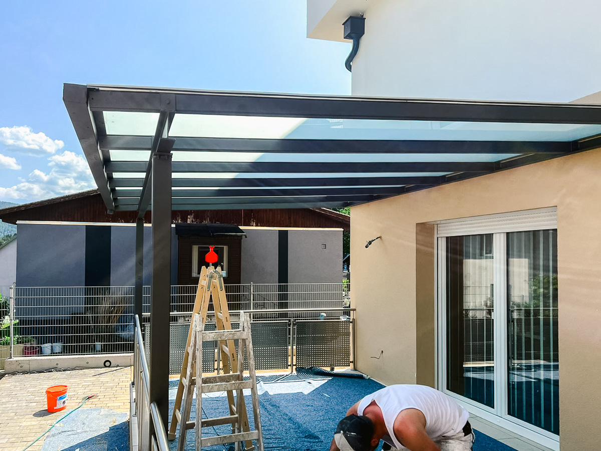 Aluminium-Terrassenüberdachung mit Glasplatten an beige verputzter Hausfassade; Maler arbeitet auf der Terrasse neben Leiter