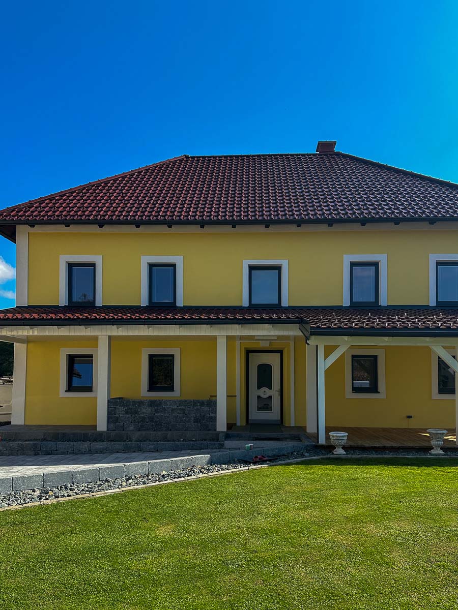 Frisch gestrichene gelbe Hausfassade mit weißen Faschen und rotem Ziegeldach, Eingangsbereich und überdachte Terrasse im Vordergrund.