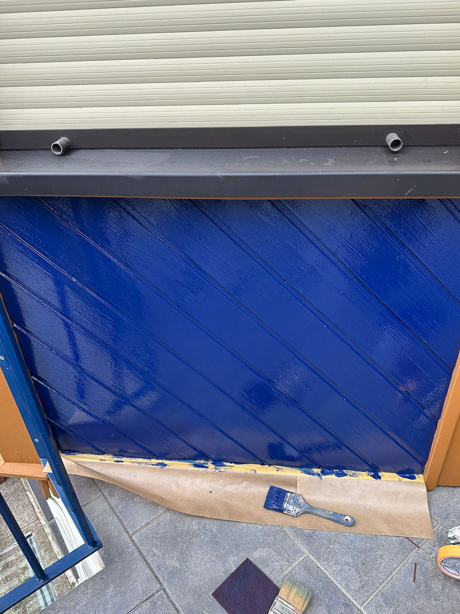 Detailaufnahme einer frisch beschichteten, dunkelblauen Holzverkleidung mit Abklebung und Malerwerkzeug auf Fliesenboden.